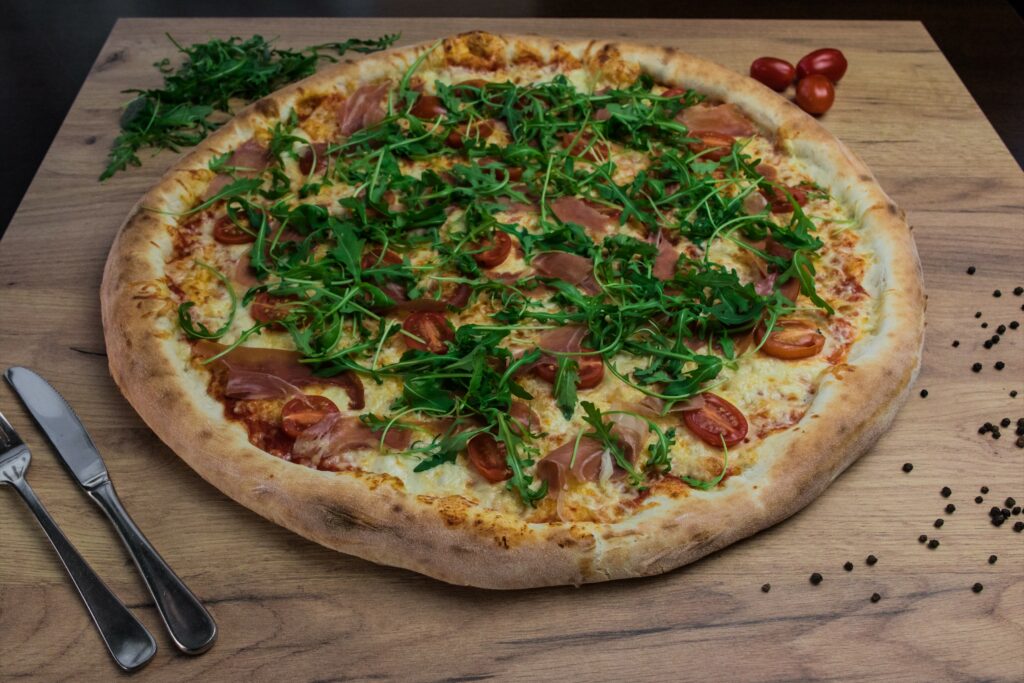 Duża pizza na drewnianej desce z rukolą, pomidorkami koktajlowymi, szynką i serem. Obok leży sztuciec.